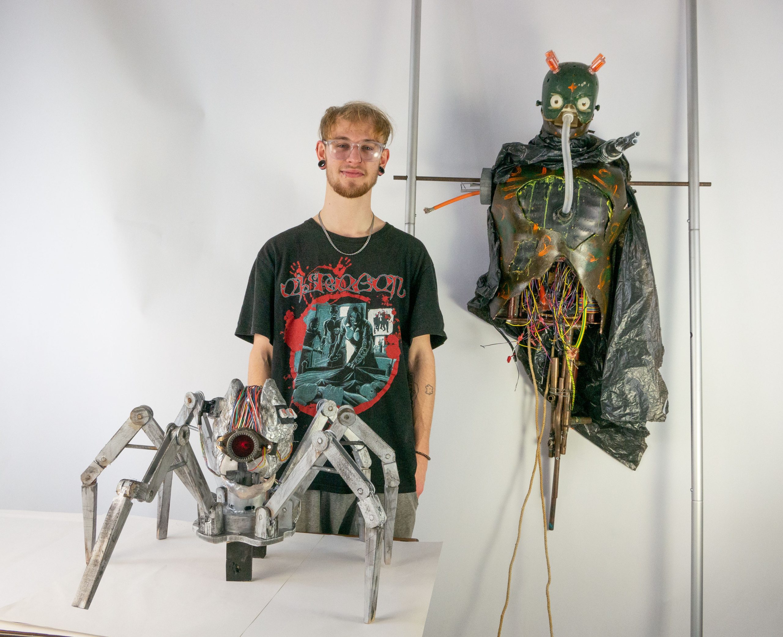Art student Osian Morris stands alsongside his work, a giant metalic spider and a disembodied robot.