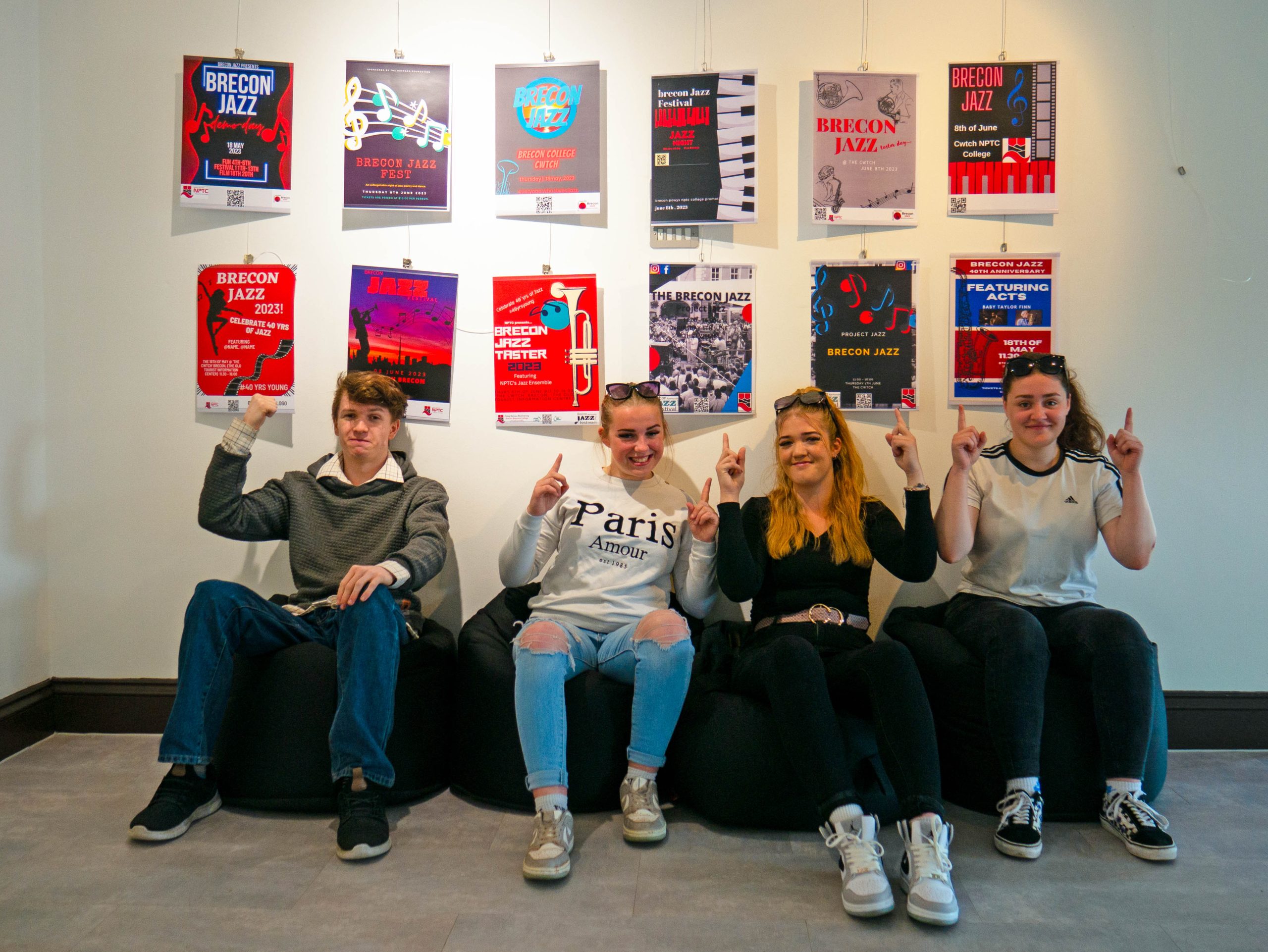 Four students sitting under Brecon Jazz Festival posters, pointing upwards at them, at the launch of the Brecon Jazz Gallery opening.