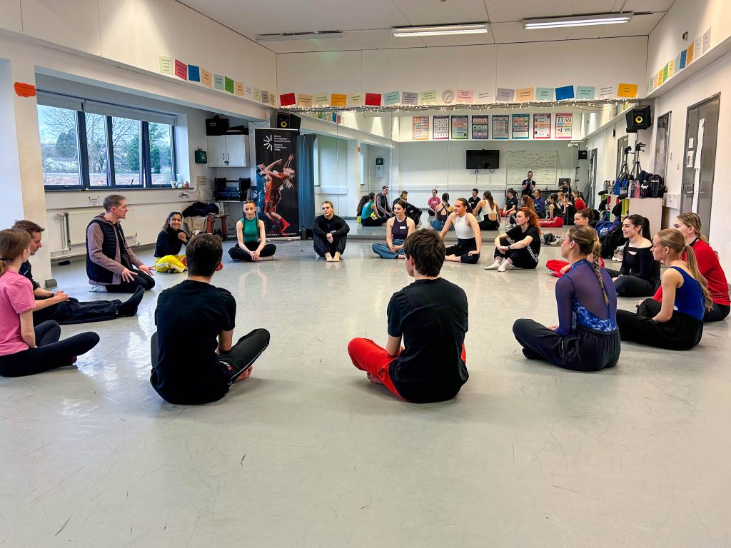 Dance group in the a dance studio sitting on the floor lostening to a lecturer